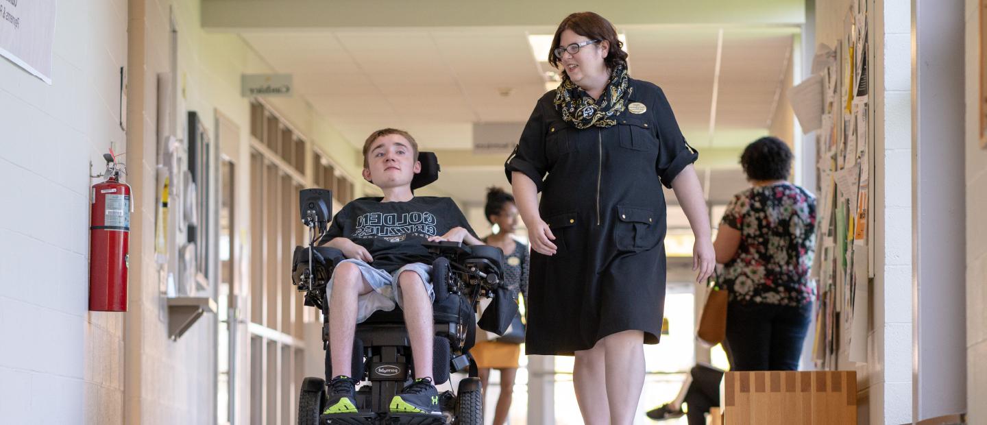 woman walking down a hallway with a young man in a wheelchair next to her