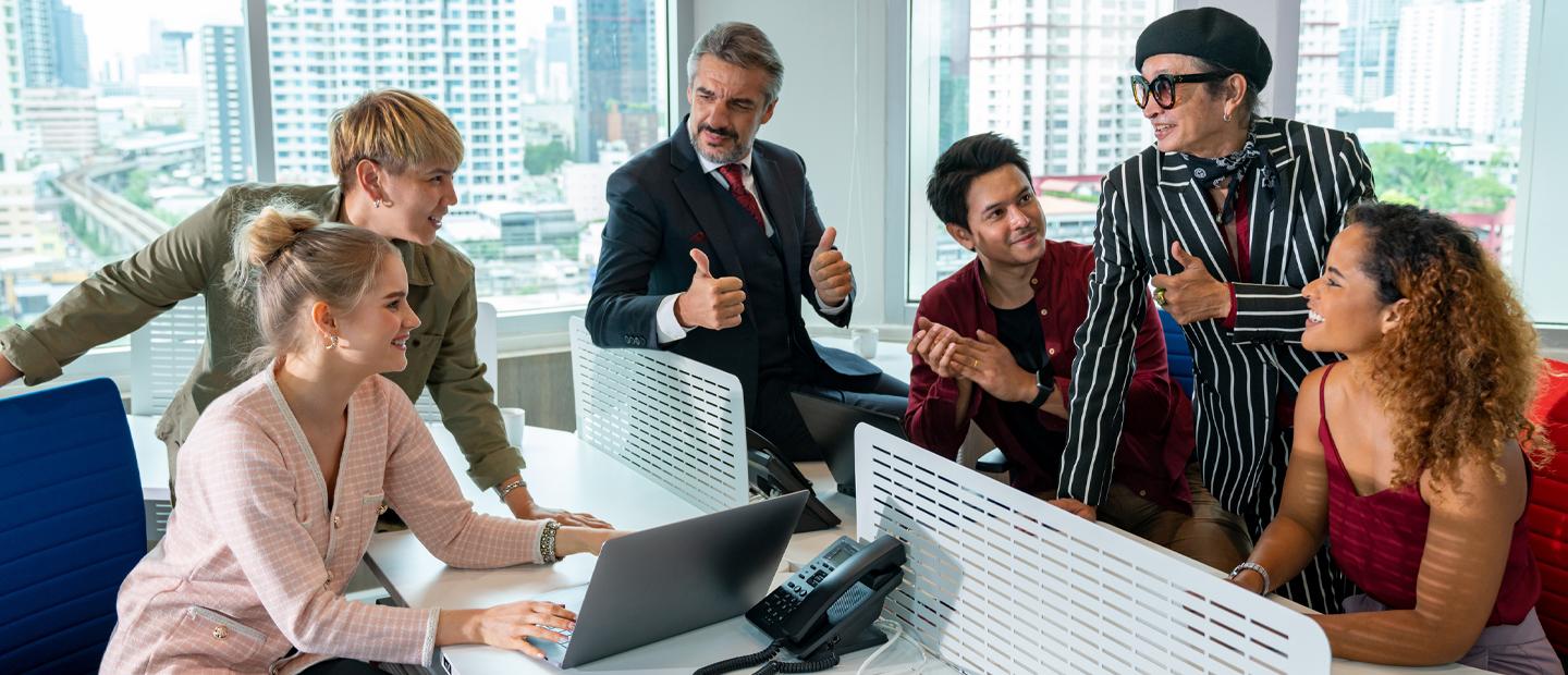 Six people sitting or standing around a desk in an office, some smiling, some giving a thumbs up signal.